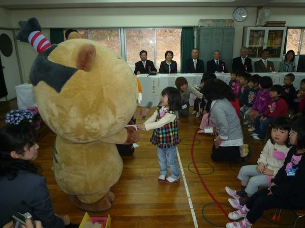 胸にピンク色の紙花をつけた園児たちが椅子に並んで座り園児の一人がマイクを向けられて、キャラクターに話をしている様子の写真